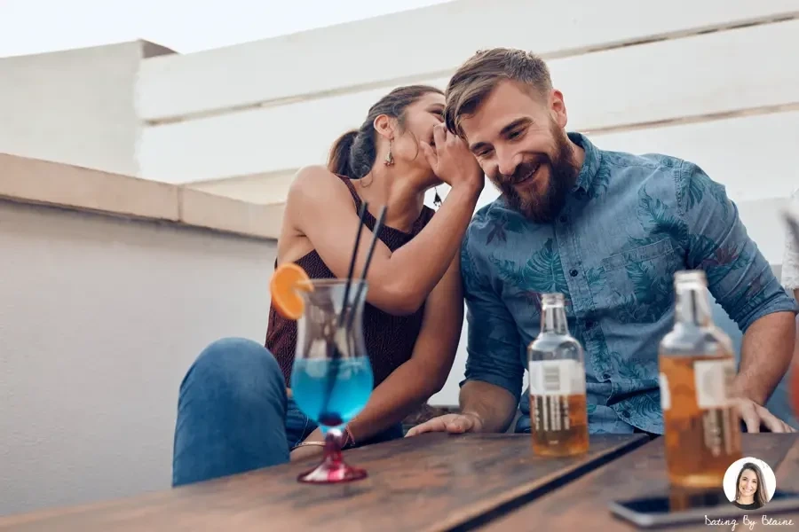 Woman and man sitting on what looks like a roof deck, at table with beers, and woman is whispering into a man's ear