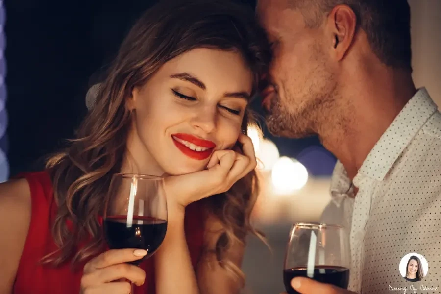 Woman and man holding glasses of wine, with woman smiling into the camera, and man whispering something in her ear