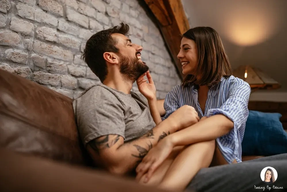 A girl putting her legs in the guys lap while touching his beard, both laughing