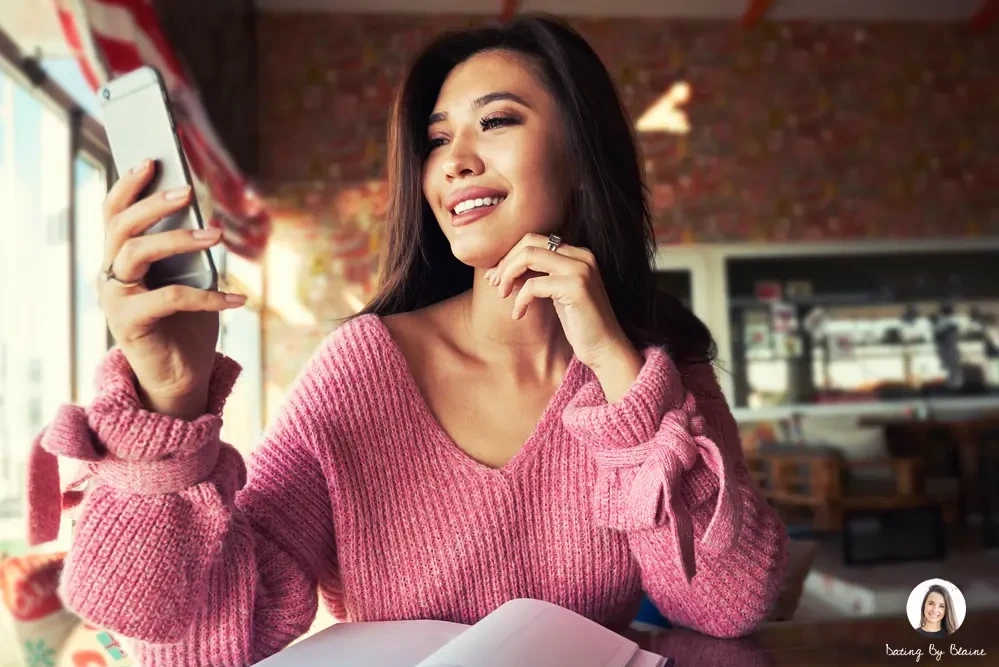 A girl in a pink sweater, holding her phone and chin, smiling