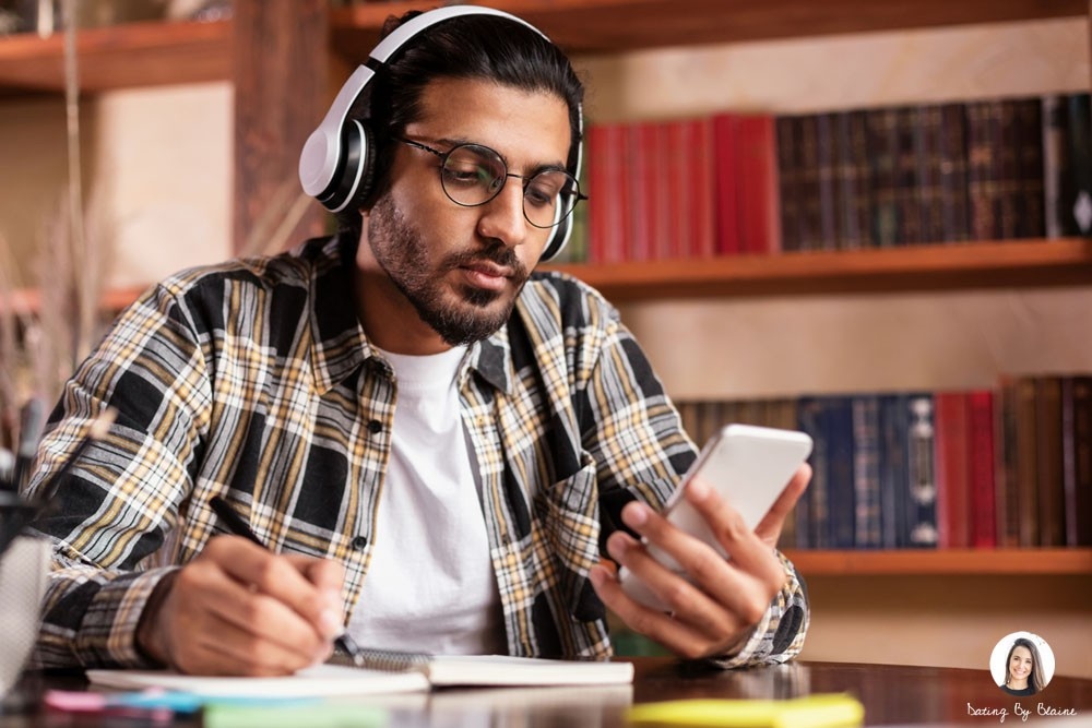 A man seated in a library, looking at his phone with one hand, taking notes with the other