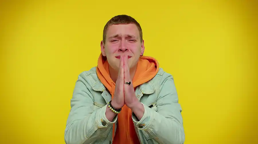 Man in an orange sweatshirt with his hands folded in front of him, looking pleading