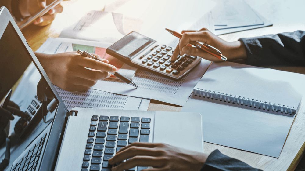 Payroll cost calculation with calculator, laptop, and financial documents showing employer of record fees, taxes, and monthly hiring expenses