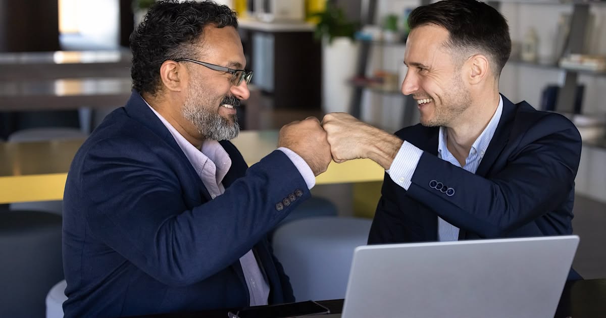 Two smiling men in navy blazers fist-bump over a laptop in a modern office.