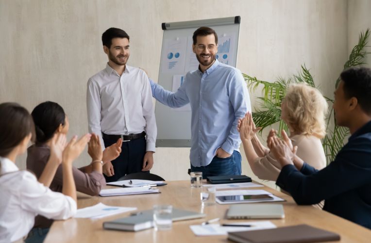 Colleagues applaud two smiling men standing by a whiteboard during a meeting in a modern office.