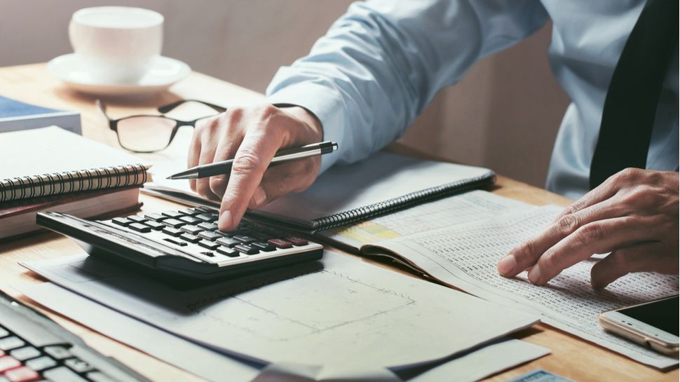 Man in a shirt and tie uses a calculator while reviewing financial documents at a desk.