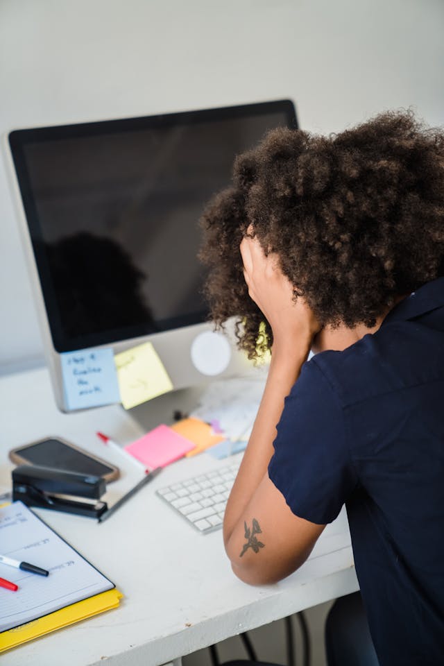 Black woman from the back, with her head in her hands at a desk with lots of sticky notes