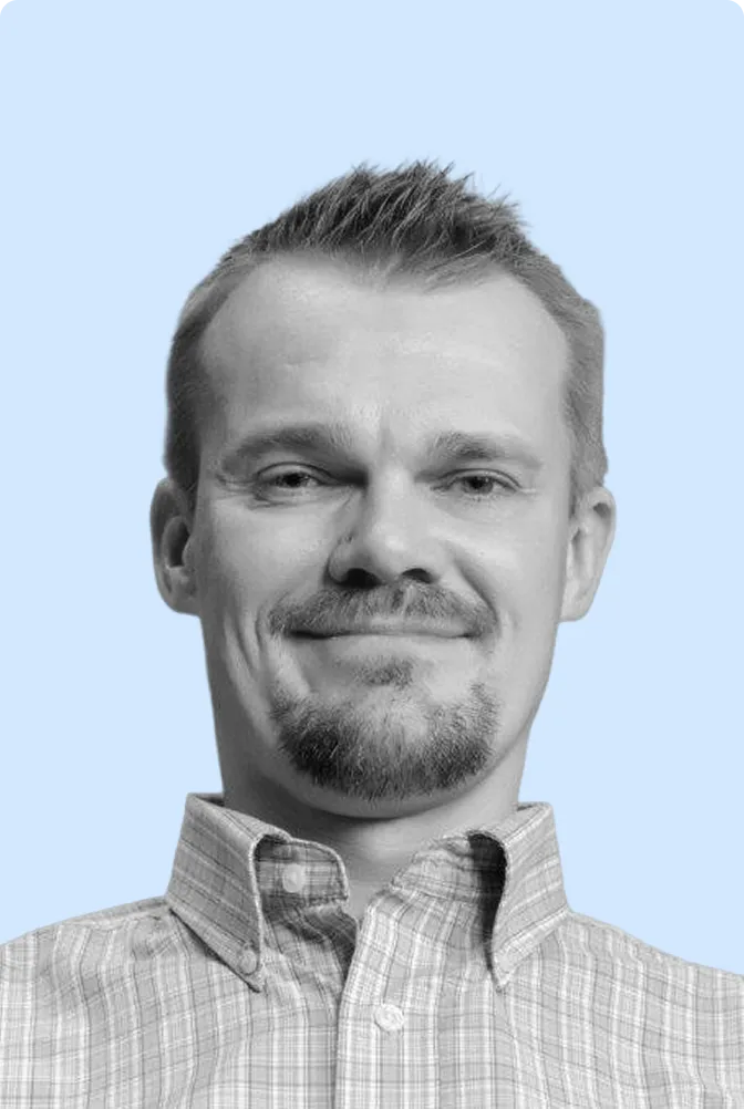 A man with short hair and a light beard, wearing a light-colored collared shirt, is smiling in front of a light blue background, evoking the relaxed vibe of surf-friendly beaches perfect for destination vacations.