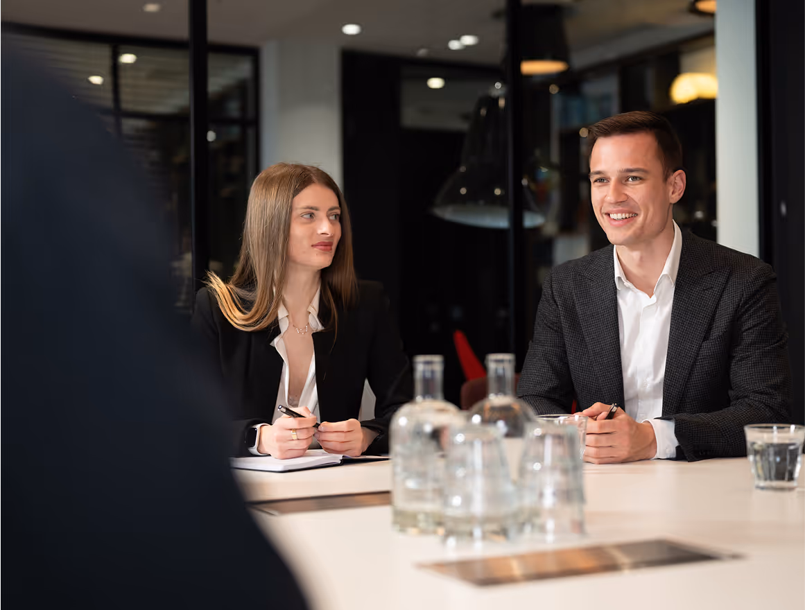 Zwei Fachleute in Geschäftskleidung sitzen an einem Konferenztisch mit Notizblöcken und Stiften, lächeln und unterhalten sich. Auf dem Tisch stehen Wasserflaschen und Gläser. Der Hintergrund zeigt eine moderne Büroumgebung.
