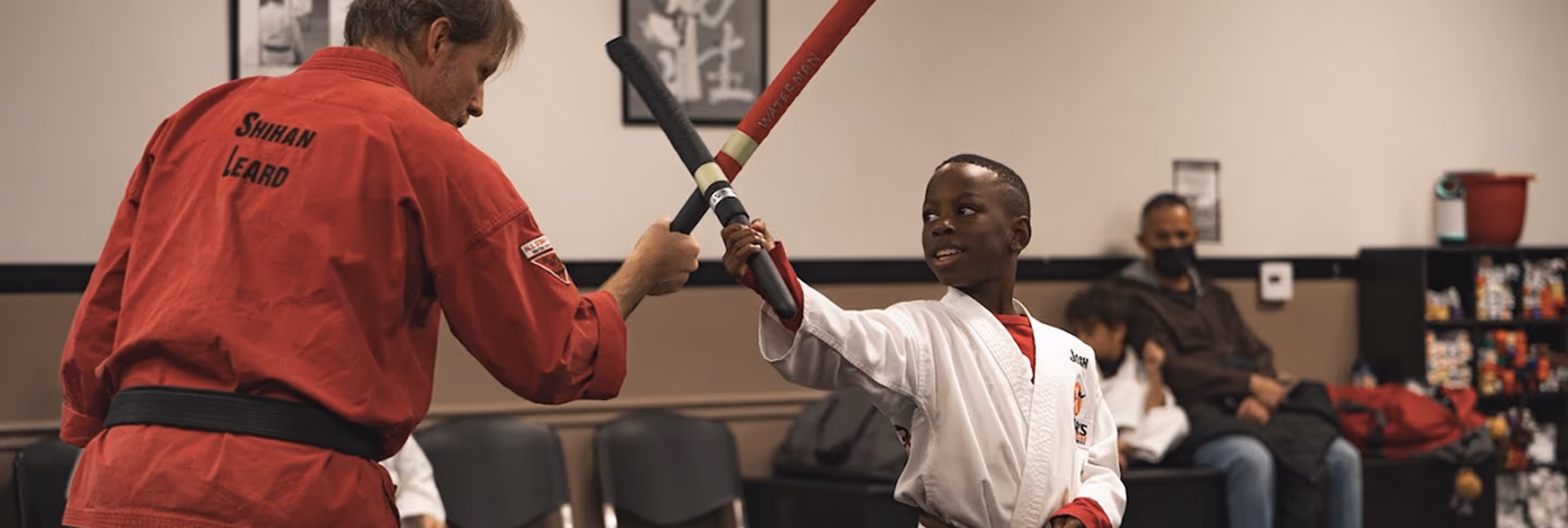 A martial arts instructor in a red uniform sparring with a boy in a white uniform using padded sticks.