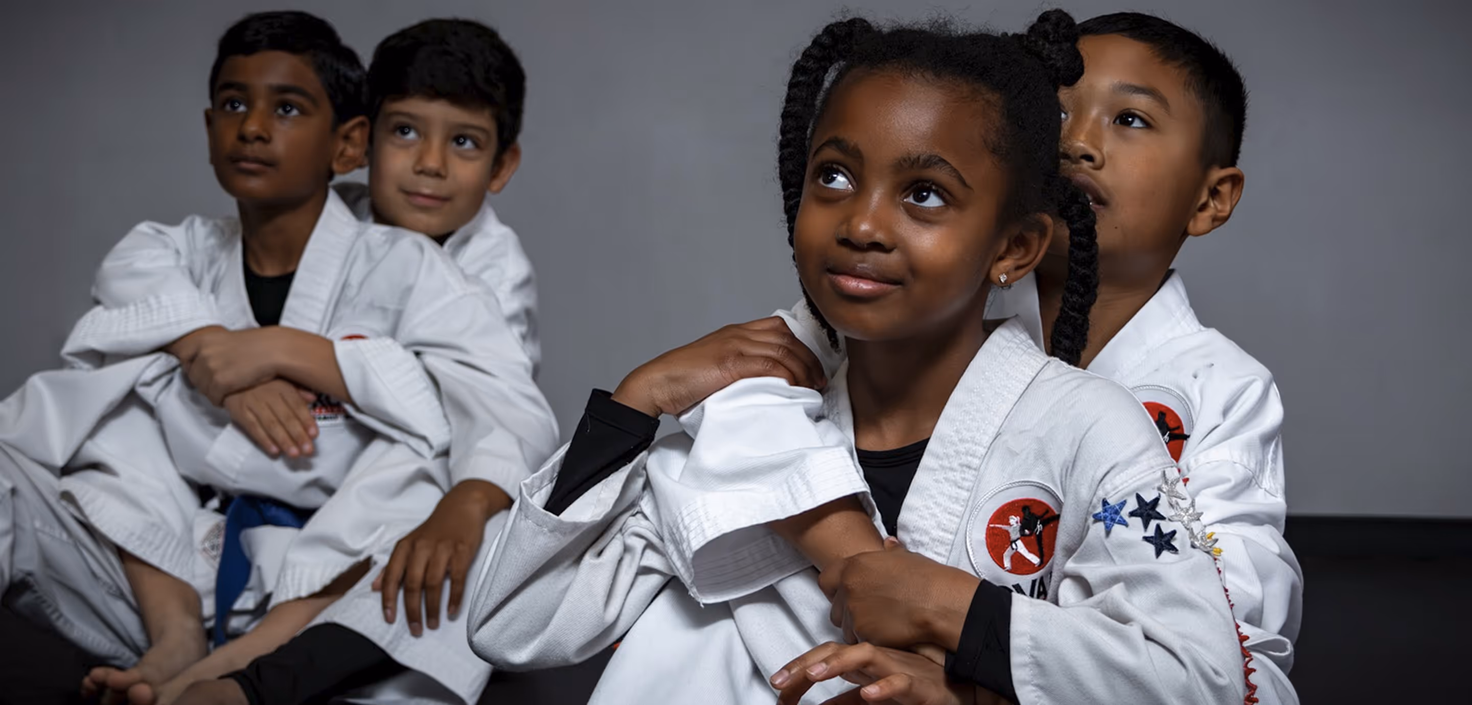 Four children in martial arts uniforms sitting together on the floor, two pairs hugging each other.