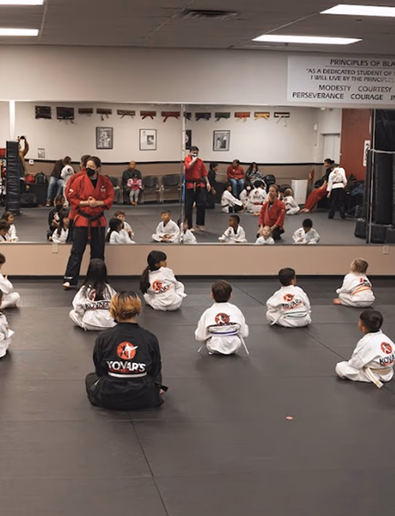 Martial arts class with children in white uniforms seated on mats facing instructors in red and black uniforms in a studio with mirrors.