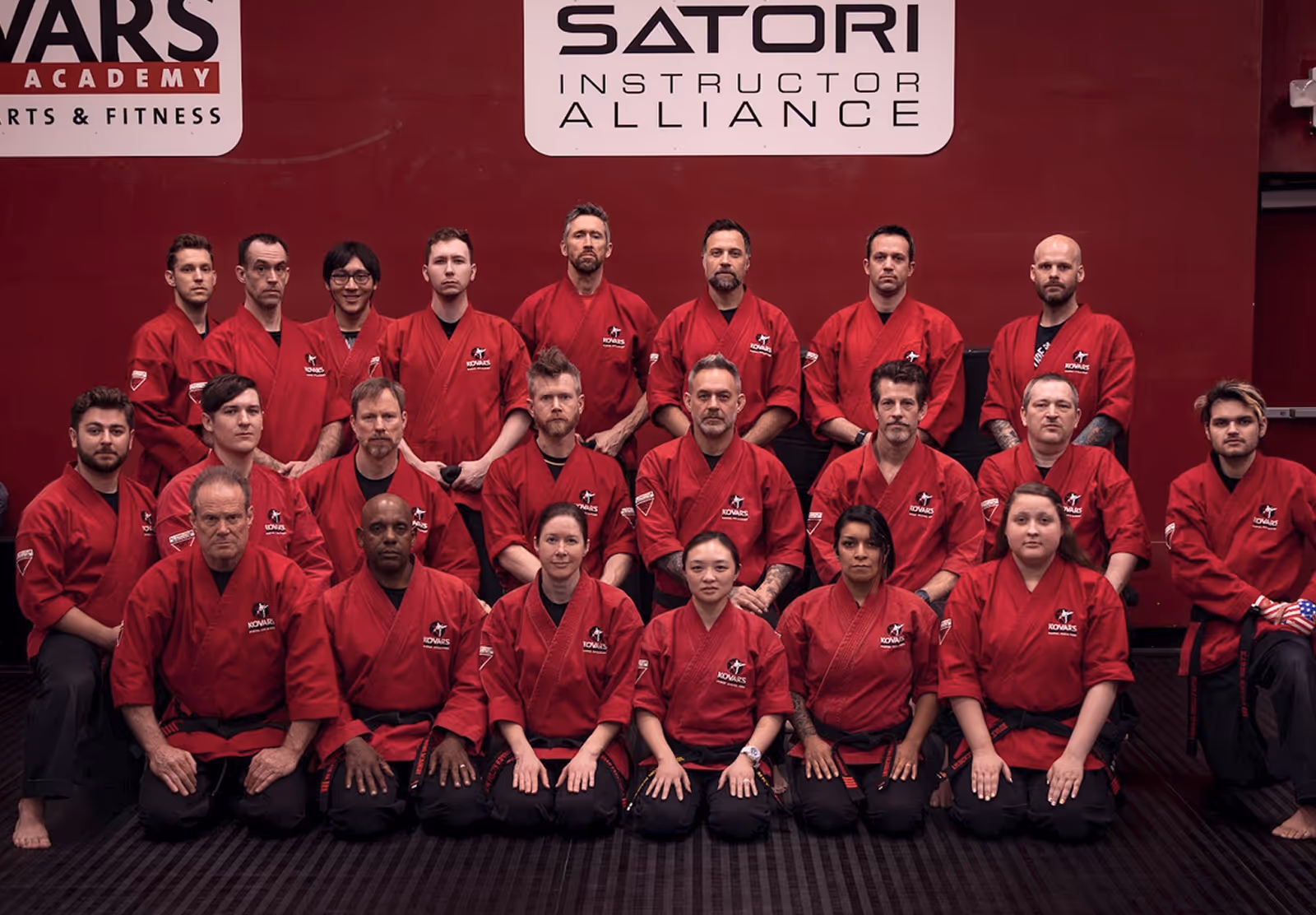 Group of martial arts students and instructors wearing red uniforms kneeling and standing in front of a maroon wall with martial arts academy logos.
