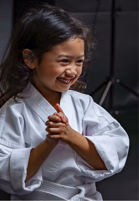 Smiling young girl in a white karate uniform with hands clasped, standing in a studio.