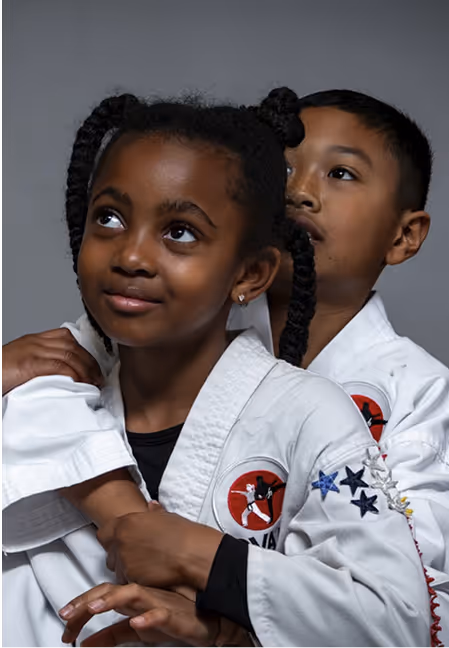 Two children in martial arts uniforms practicing a hold, with the boy standing behind the girl.