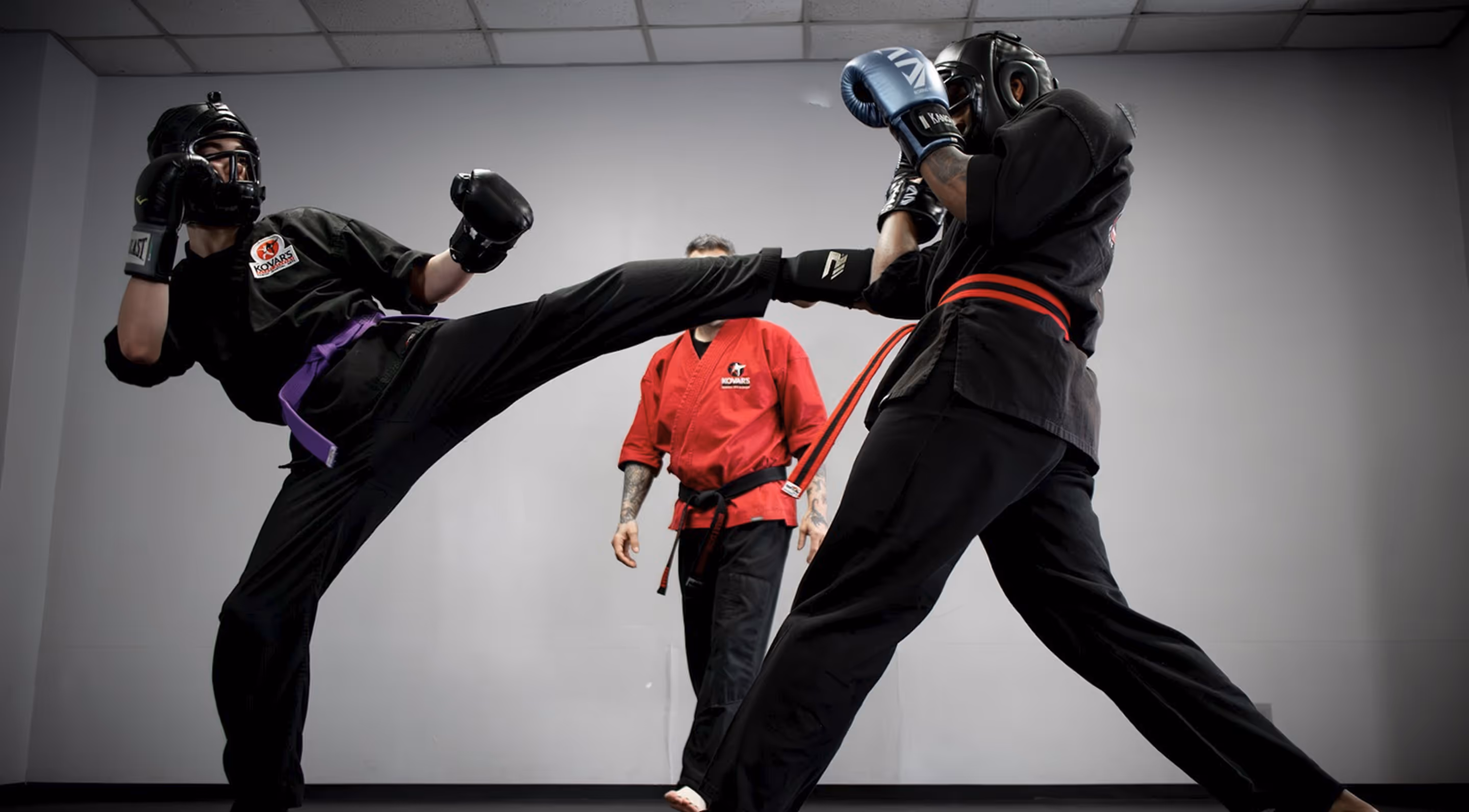 Two martial artists sparring indoors wearing protective headgear and gloves, with one delivering a high kick and an instructor in a red uniform observing in the background.