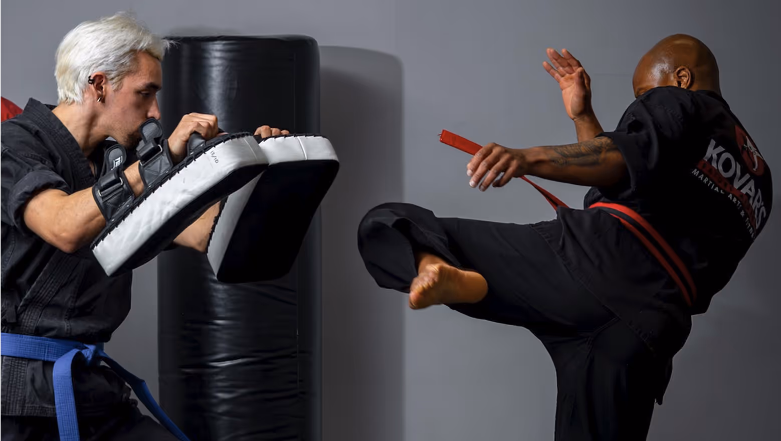 Martial artist with a red belt executing a high kick towards pads held by a second martial artist in a blue belt.