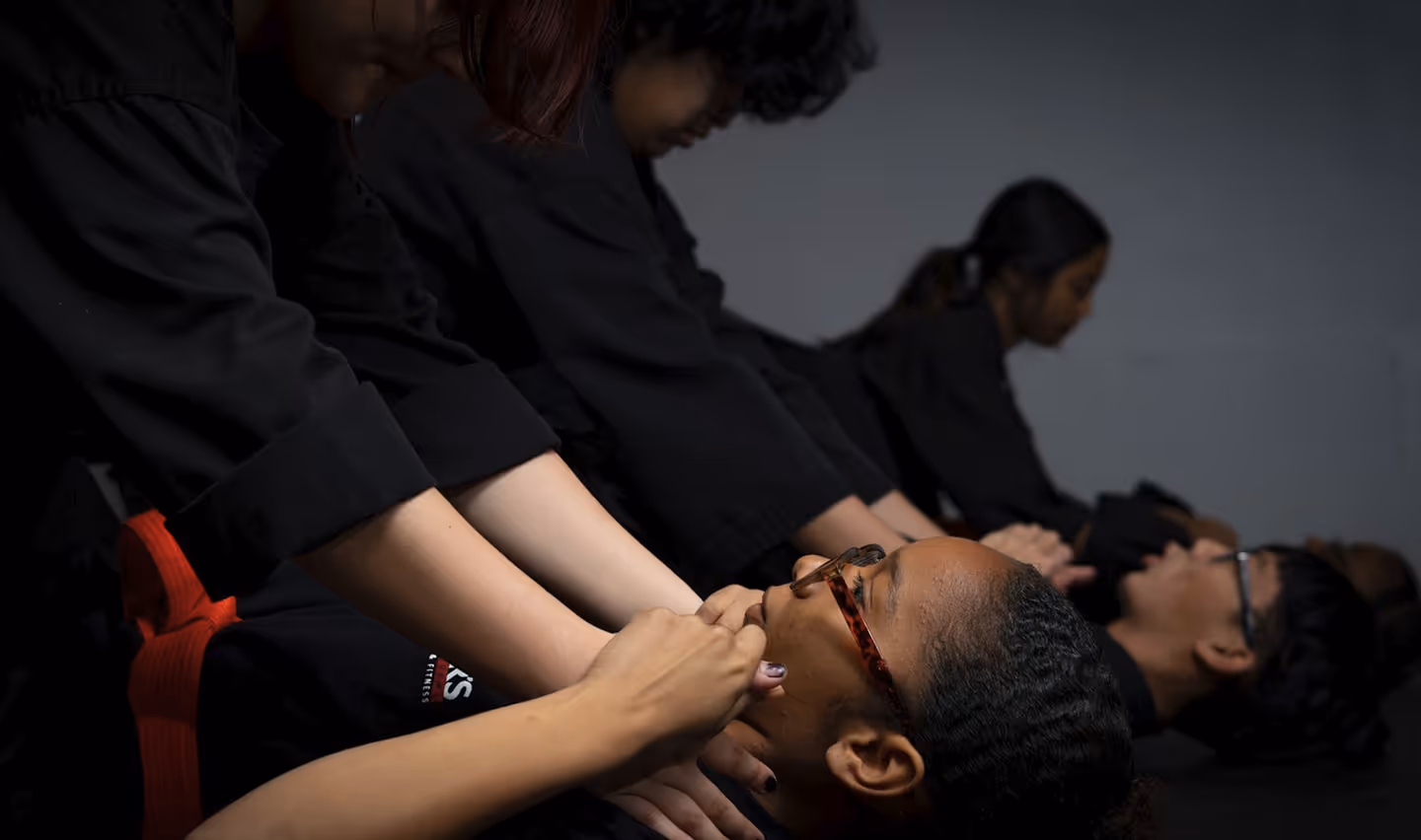 Group of people in black clothing practicing a self-defense technique on a person lying down with glasses.