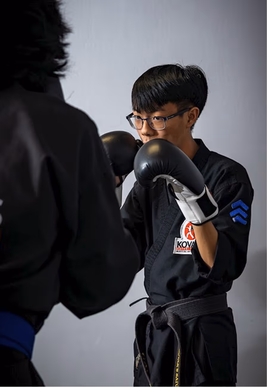 Young martial artist wearing glasses and black uniform with black belt and gloves in fighting stance facing an opponent.