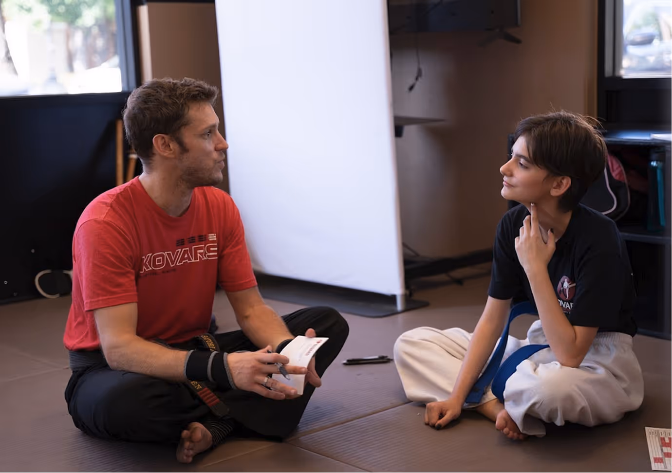 Martial arts instructor and young student sitting cross-legged on a mat, engaging in discussion during a class.