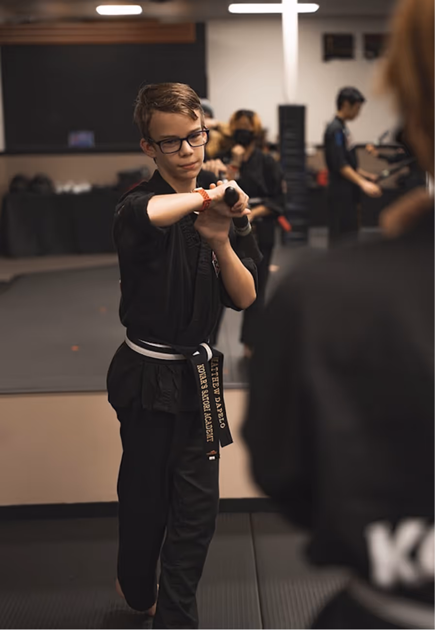 Young martial arts student wearing glasses and a black belt practicing with a training stick in a dojo.
