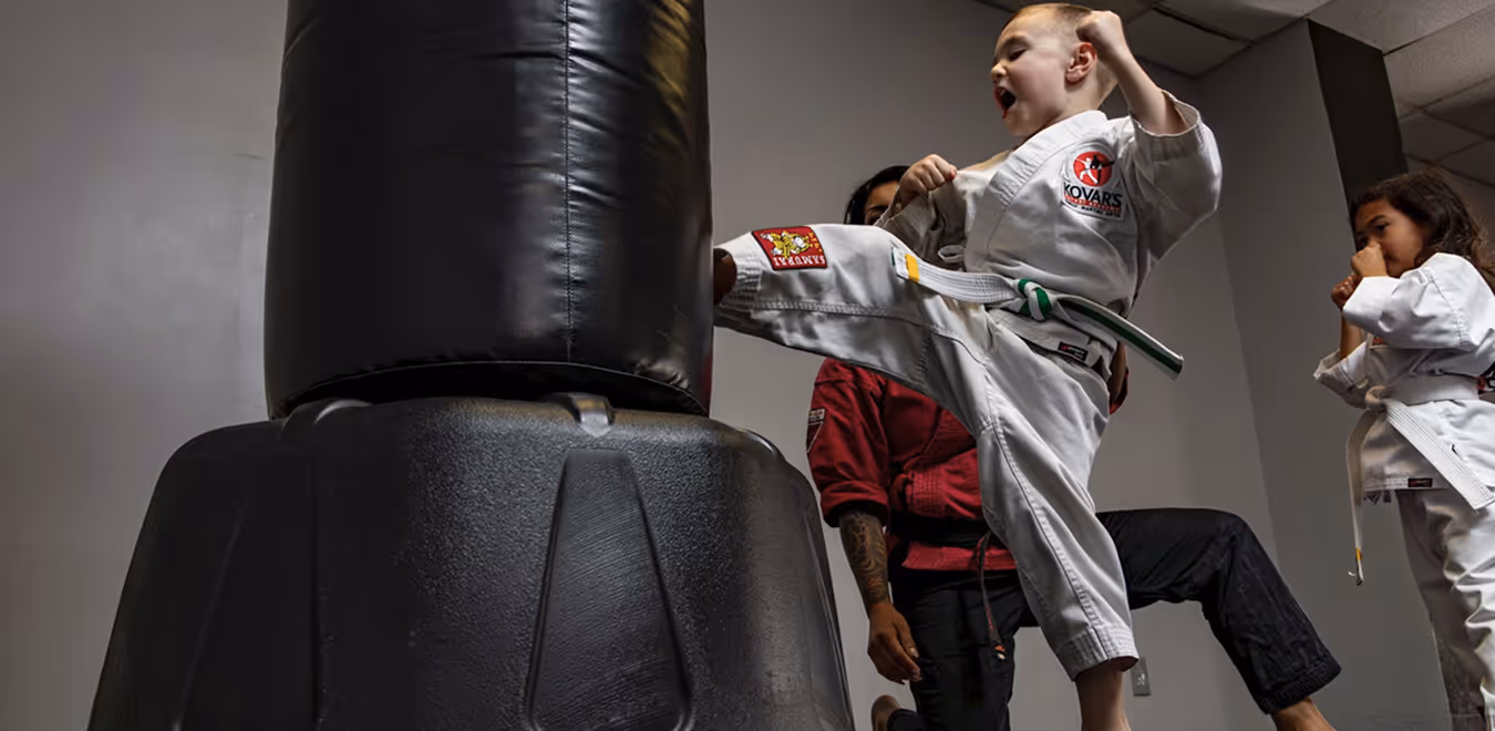 Young martial arts student in white gi with green belt kicking a black heavy bag, while an instructor and another child watch.