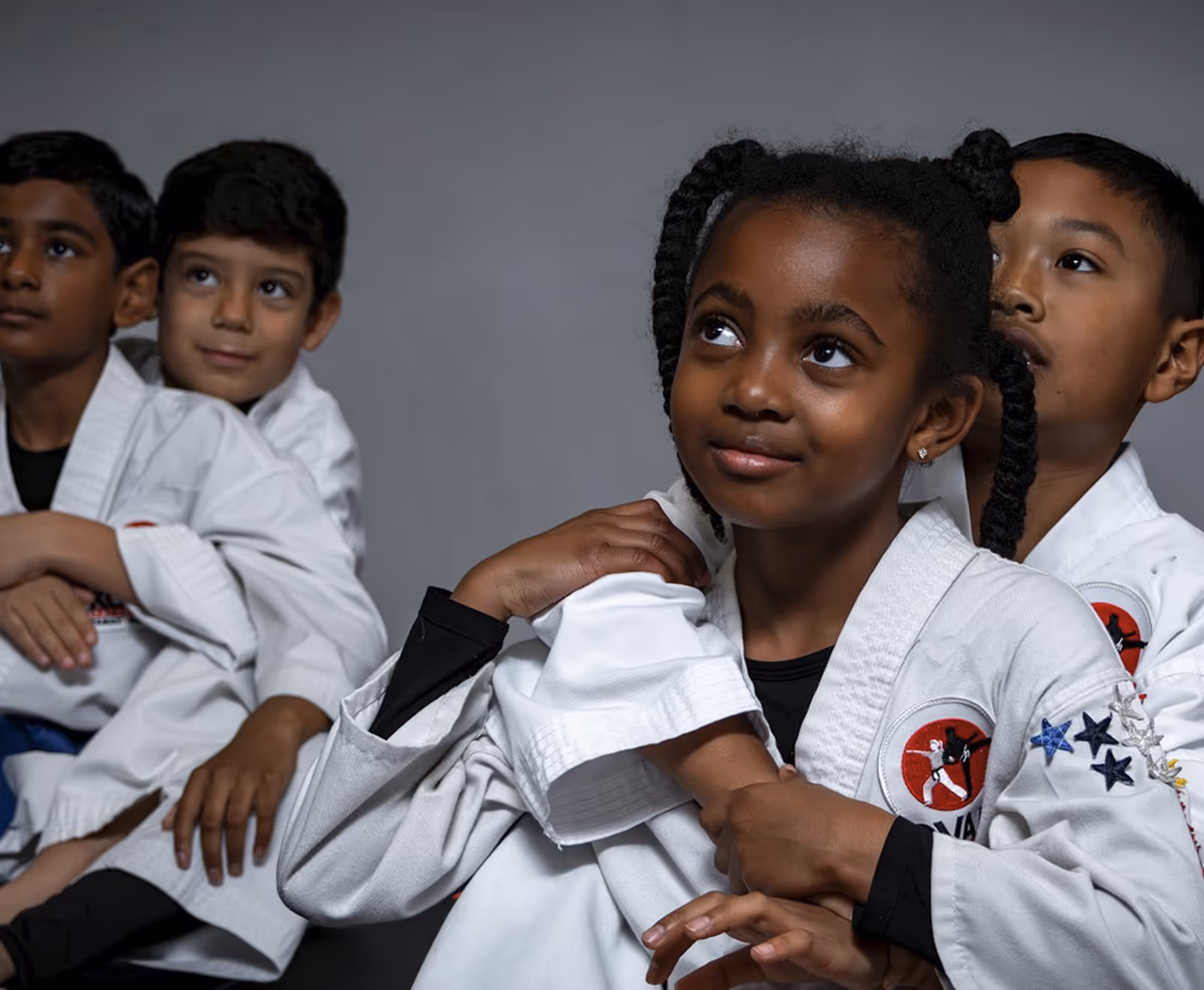 Four children in martial arts uniforms sitting closely together against a plain background.