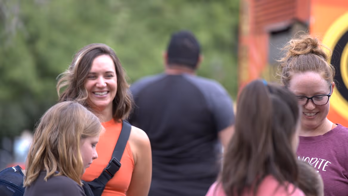 Group of people, including two smiling women facing young girls outdoors with greenery in the background.