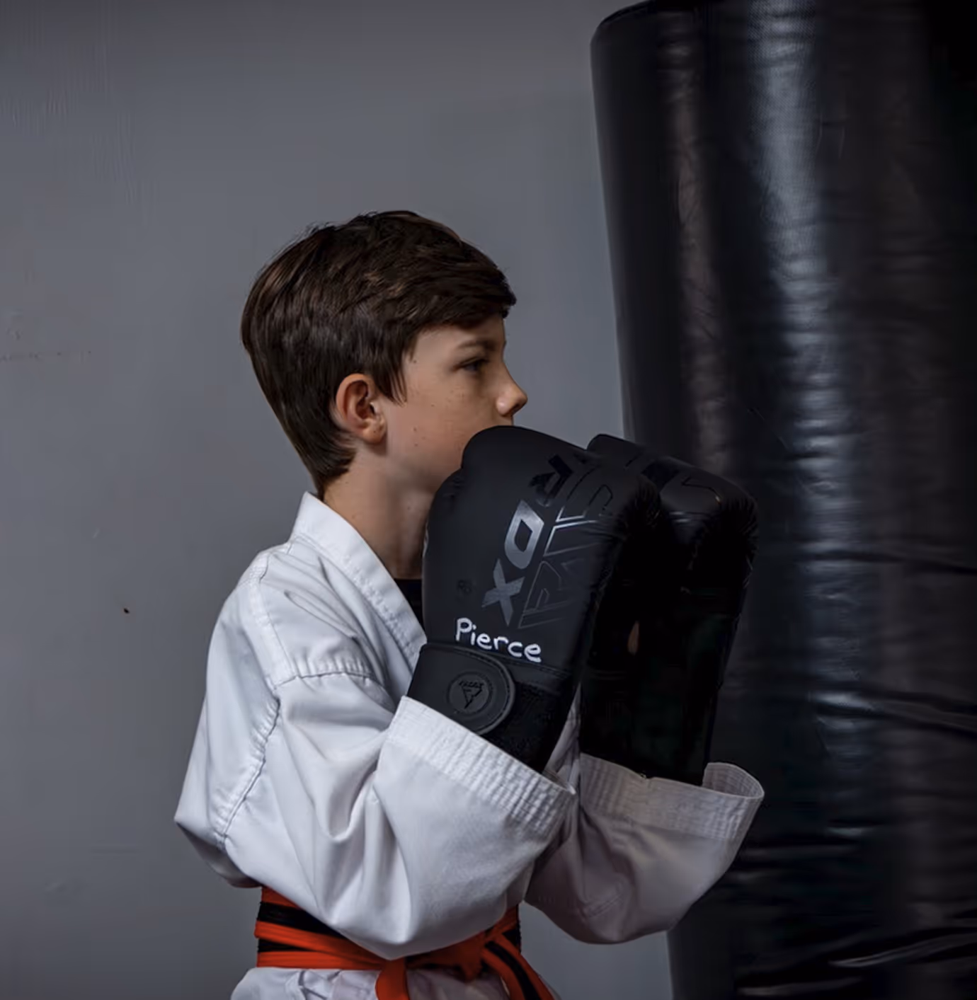 Young boy in a white martial arts uniform with a red and black belt wearing black boxing gloves, facing a black punching bag.