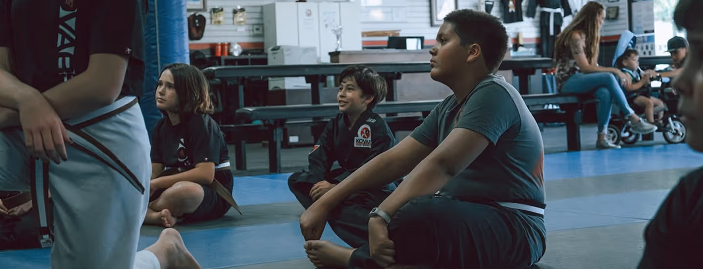 Children sitting on mats in a martial arts class, listening attentively with some wearing uniforms and belts.