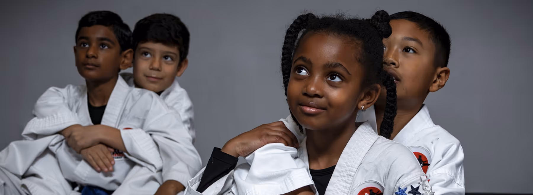 Four children in martial arts uniforms sitting in pairs, two girls and two boys, looking upward with thoughtful expressions.