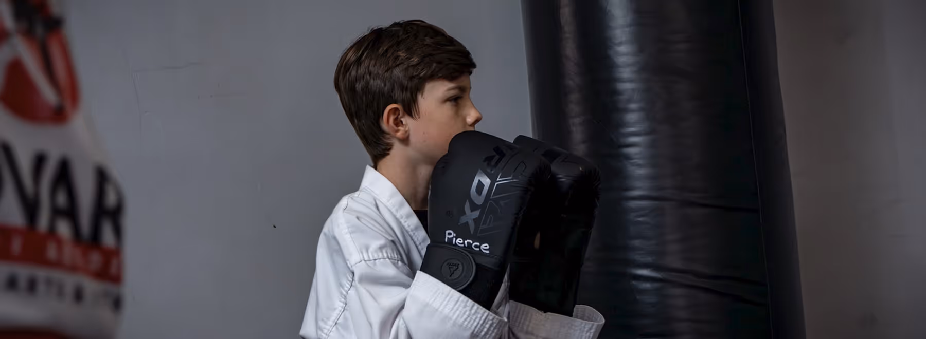 Boy in white martial arts uniform wearing black boxing gloves standing in front of a punching bag.