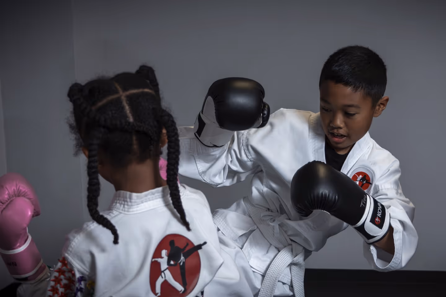 Two children practicing martial arts wearing white gi uniforms and boxing gloves in a training room.