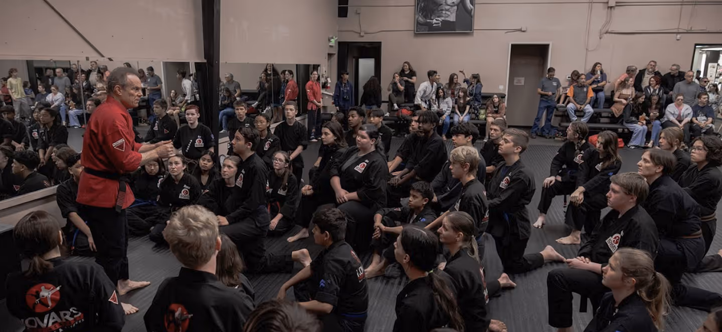 Martial arts instructor in a red uniform teaching a large group of students dressed in black uniforms seated on the floor in a dojo.