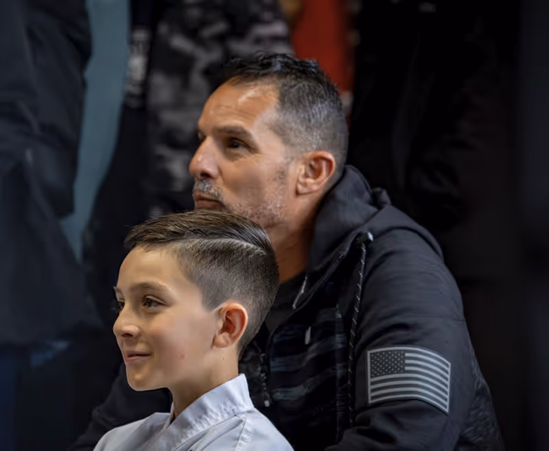 Portrait of a boy in a white martial arts uniform with a man in a black hoodie featuring an American flag patch on the sleeve in the background.