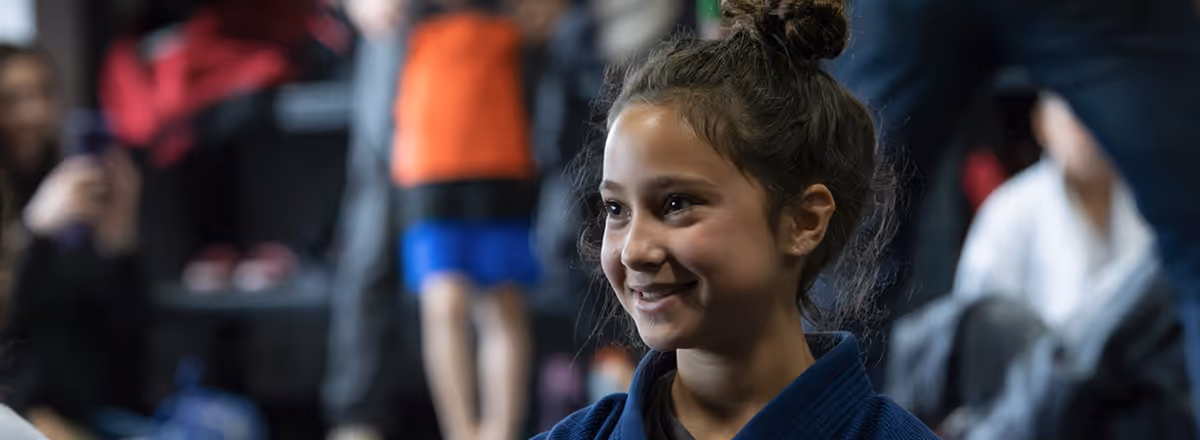 Smiling young girl in a navy blue martial arts uniform with a bun hairstyle, with blurred people and gym background.