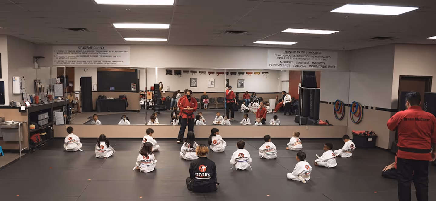 Children in white martial arts uniforms seated on a dojo floor facing an instructor in a red uniform with a black belt.