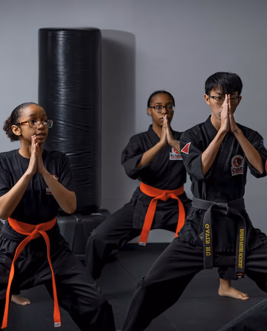 Three martial artists in black uniforms with orange and black belts performing a focused prayer pose in a dojo.