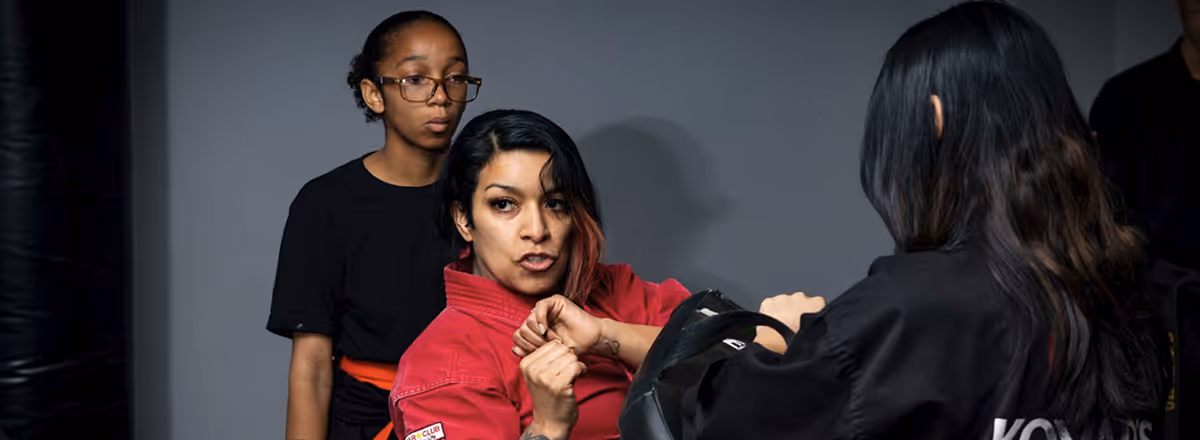 Three women practicing martial arts indoors, one wearing a red gi with focused expression, another holding a padded shield, and a third watching in the background.