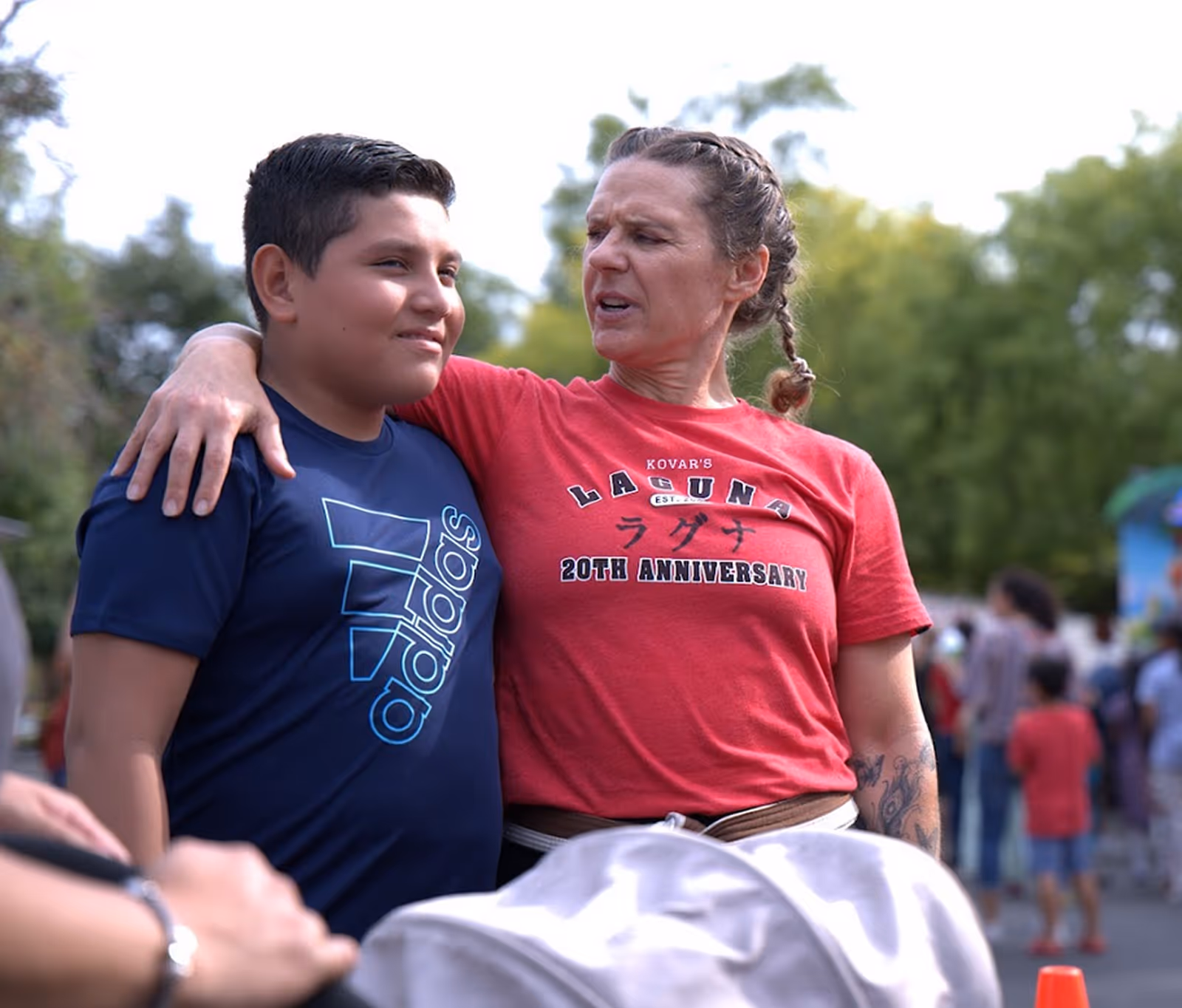 A woman in a red Laguna 20th Anniversary t-shirt with her arm around a smiling boy in a blue Adidas t-shirt outdoors.