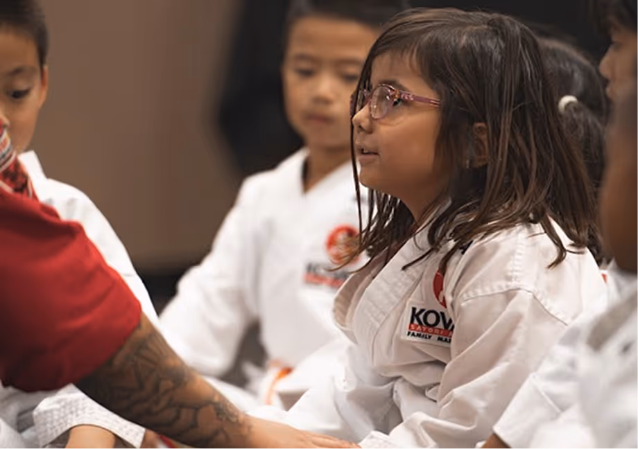 Children in white martial arts uniforms sitting and interacting during a class, with one girl wearing glasses in focus.