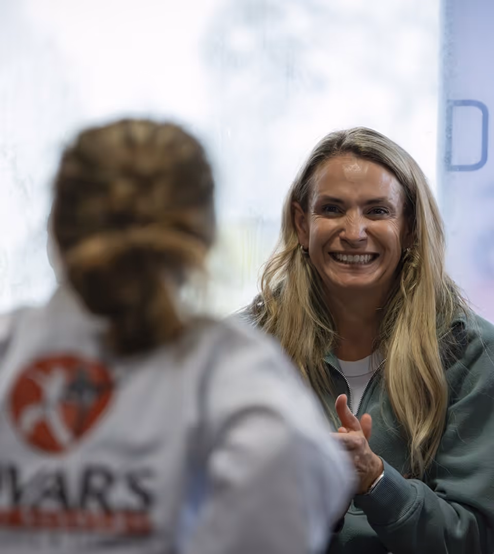 Smiling woman with long blonde hair in a green sweatshirt engaging in conversation with a blurred person in the foreground.
