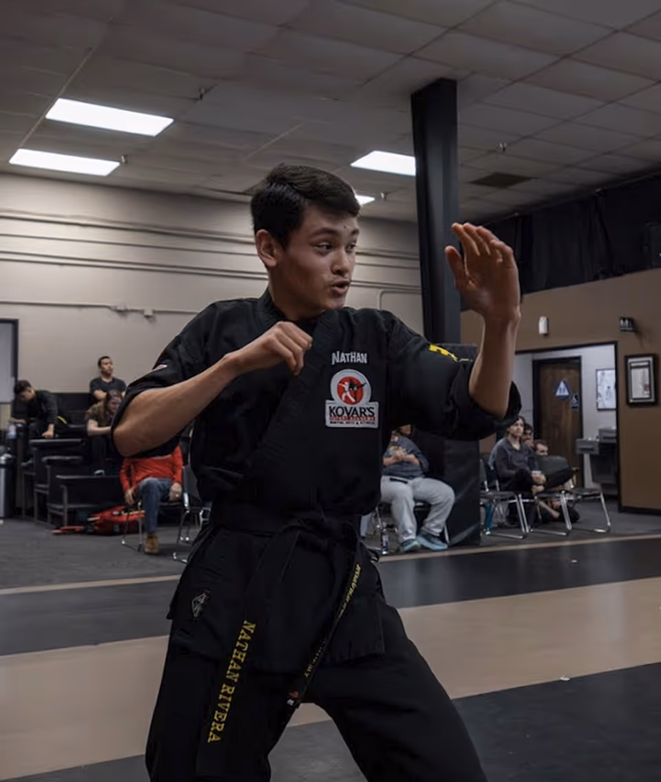 Young martial artist in black uniform practicing a defensive stance inside a dojo with seated spectators in the background.