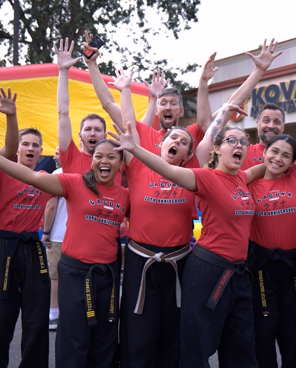 Group of smiling martial artists in red shirts and black belts celebrating outdoors with raised hands.
