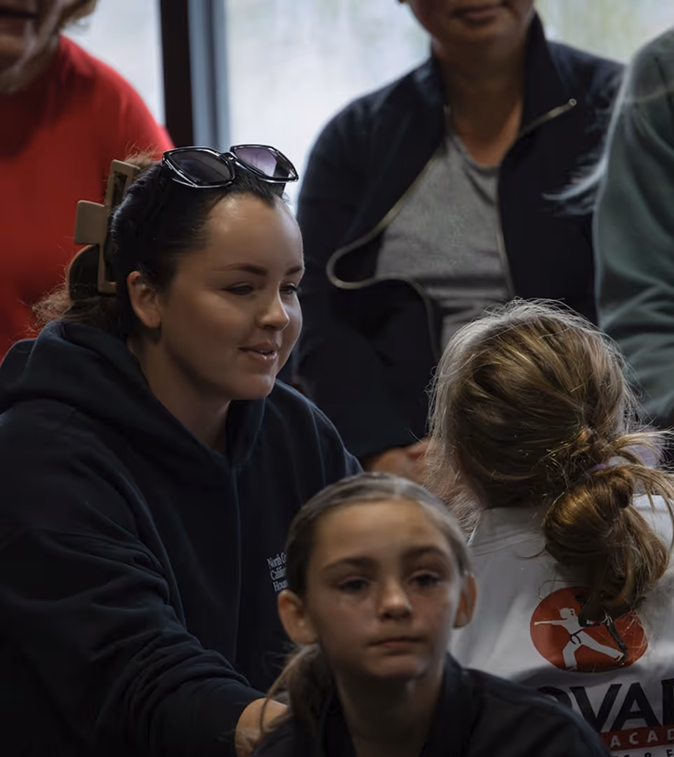 A group of people including a woman with sunglasses on her head and two girls, one wearing a jacket with a fencing logo on the back.