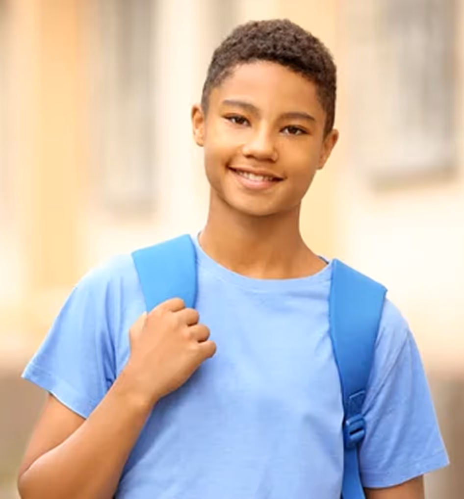Smiling teenage boy with short curly hair wearing a light blue shirt and blue backpack outdoors.