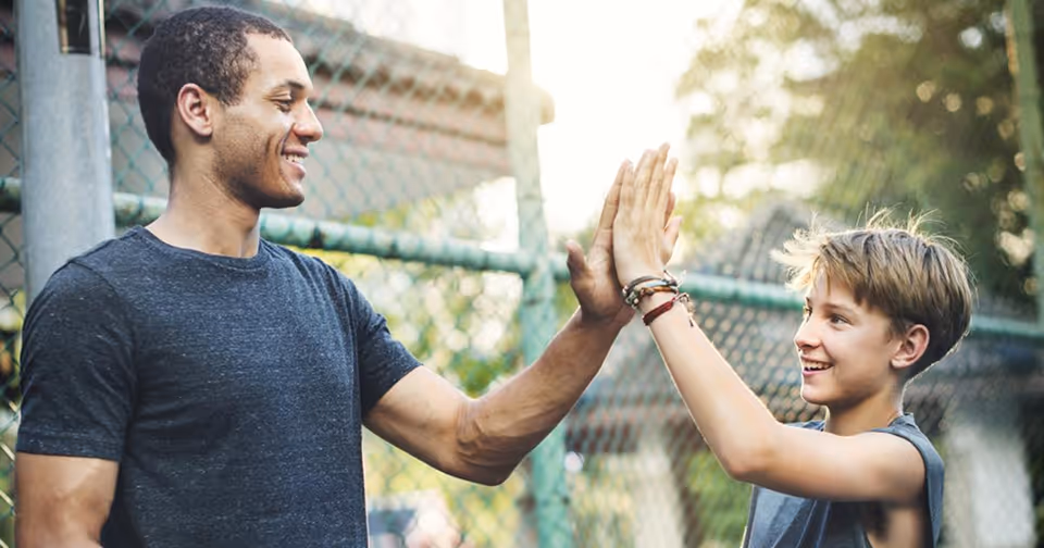 Smiling adult and young boy giving each other a high five outdoors near a chain-link fence.
