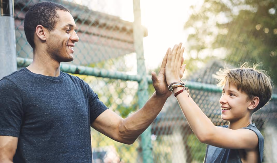 Smiling man and boy giving each other a high five outdoors near a chain-link fence.