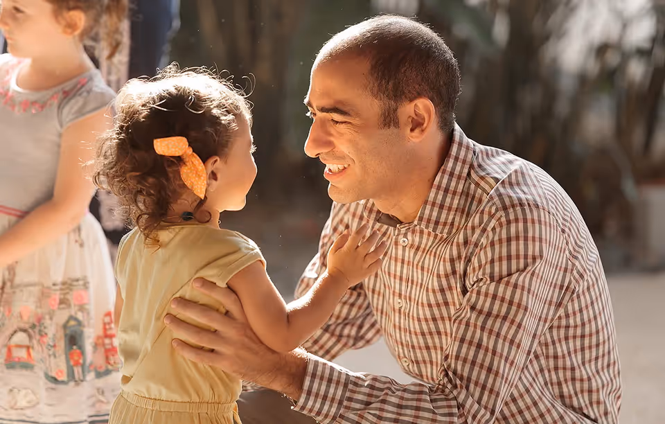 Man smiling and holding a toddler girl with a bow in her curly hair while another child stands nearby.