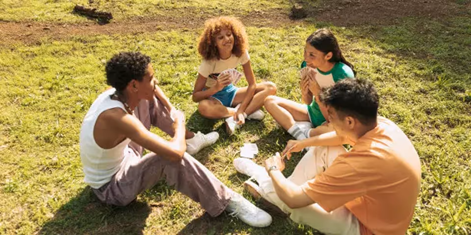 Four diverse children sitting on grass in a circle playing a card game outdoors in sunlight.